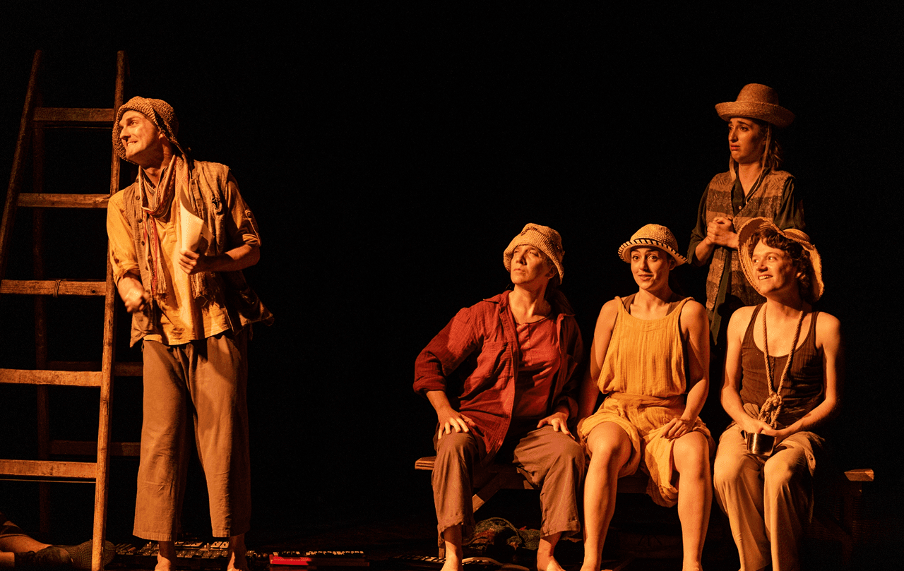 Actors performing in Flute Theatre’s adaptation of A Midsummer Night’s Dream. Five performers in rustic costumes and hats are seated or standing on stage in warm theatrical lighting, engaging with the audience in an inclusive Shakespeare performance.
