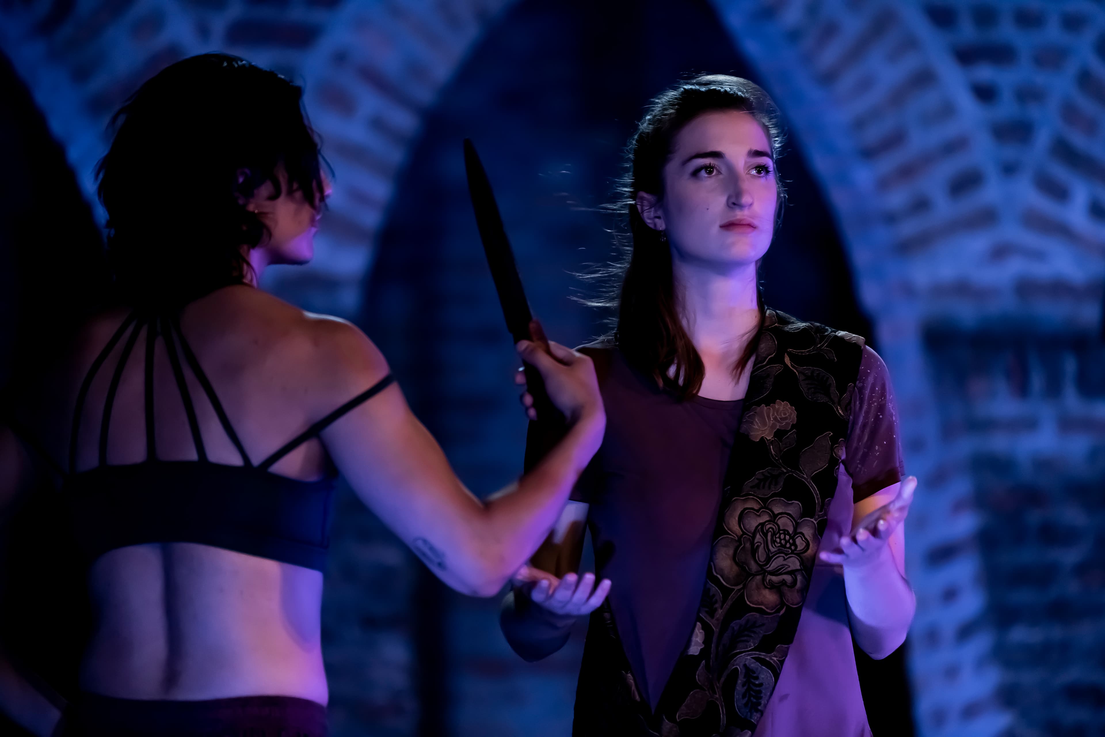 Two performers in dramatic lighting during a theatrical scene; one holds a dagger to the other, who remains calm and composed under a blue-lit stone arch backdrop.