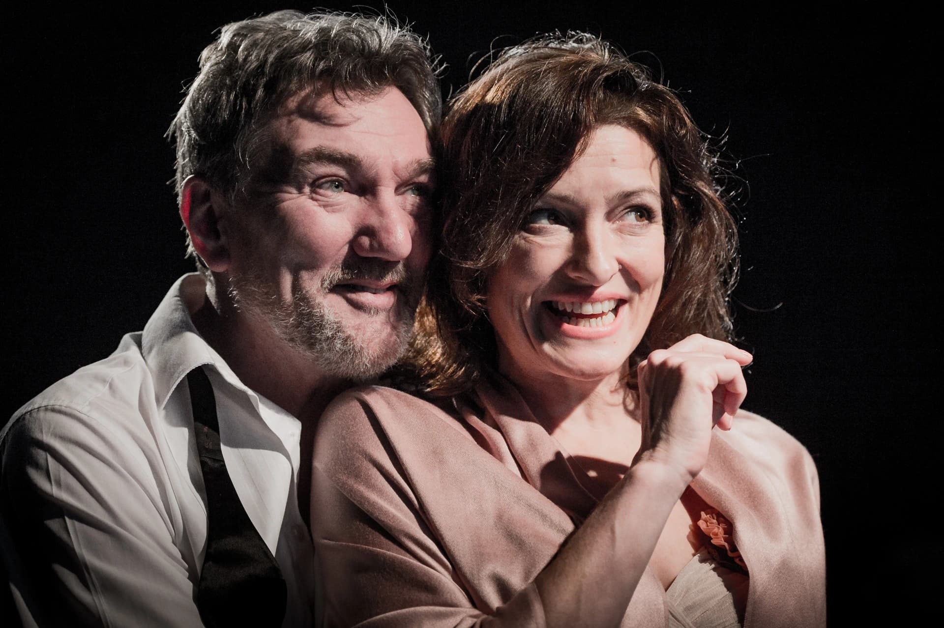 A close-up from the Hamlet main stage show (not for autistic audiences), featuring two actors in an intimate, tense moment. The man, wearing a white shirt and black tie draped loosely, smiles softly, while the woman, in a light pink wrap, has an expressive, slightly nervous smile under dramatic stage lighting.