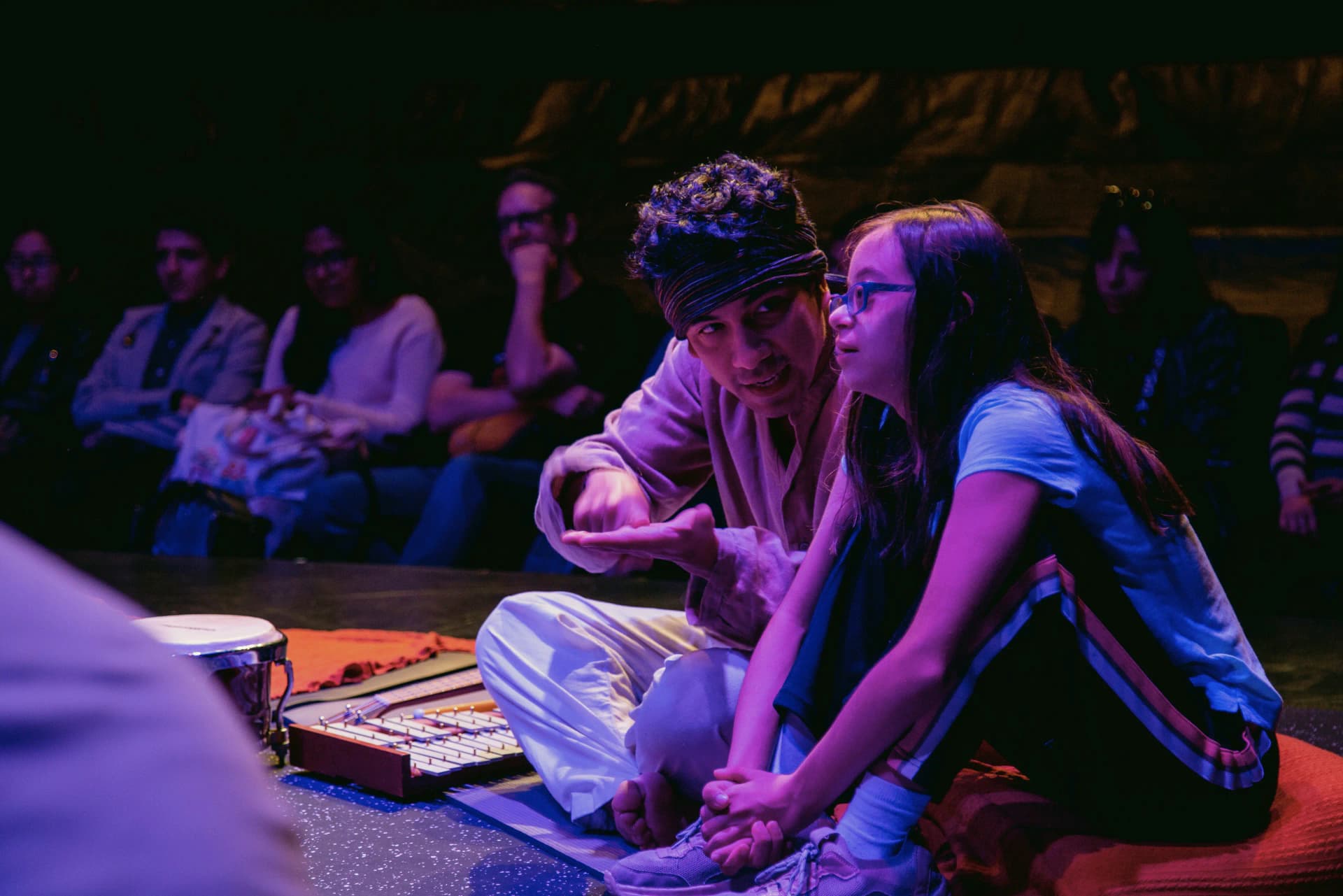 A performer sits beside a young girl in an intimate theatre setting, engaging her in a calm and focused interaction while surrounded by percussion instruments.