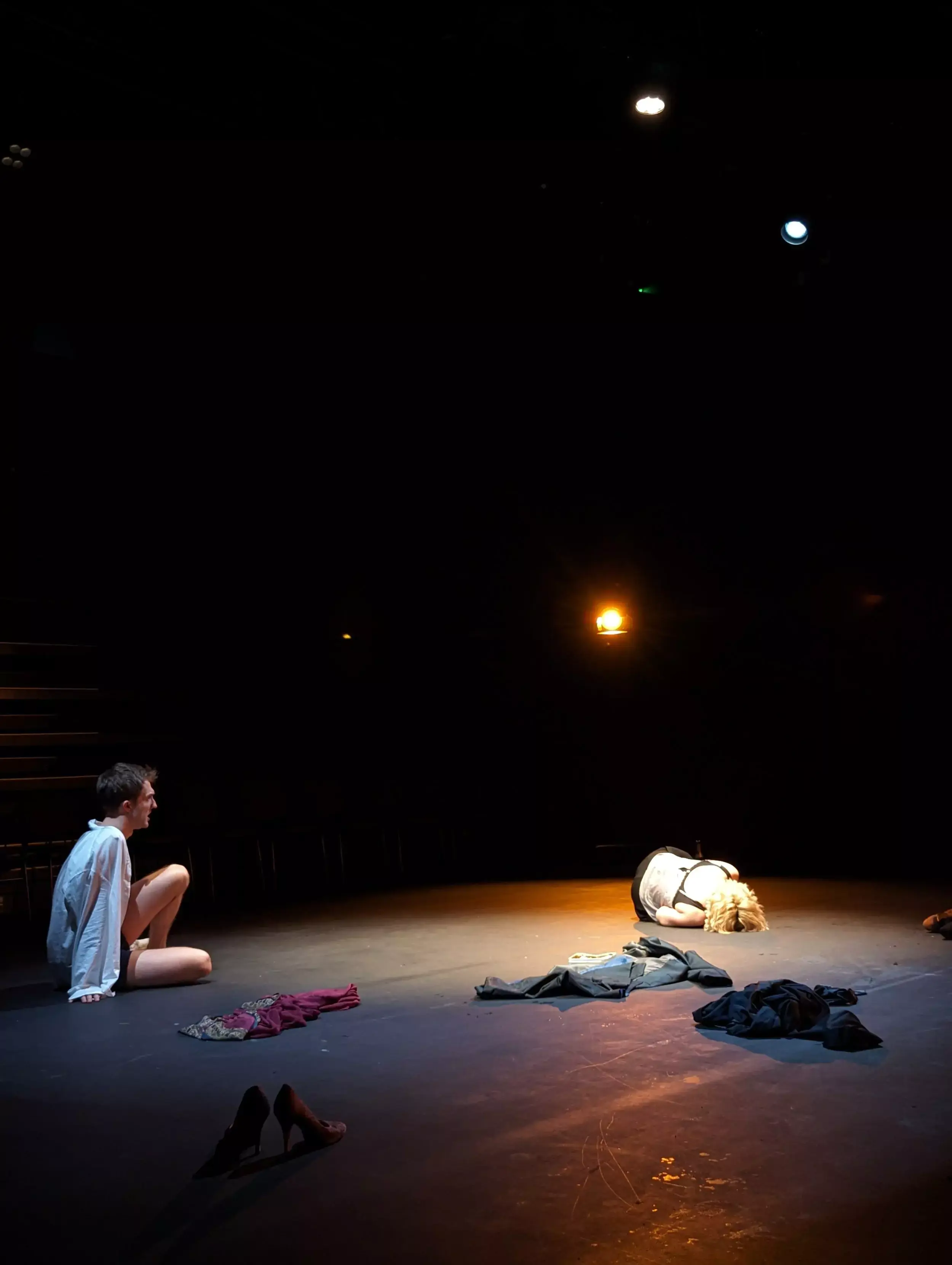 A dark, minimal stage scene from Hamlet: Collateral Damage, with two actors under soft spotlights; one sits on the floor in a white shirt, while the other curls on the ground in despair, surrounded by scattered clothing and shoes, evoking a raw and intimate moment.