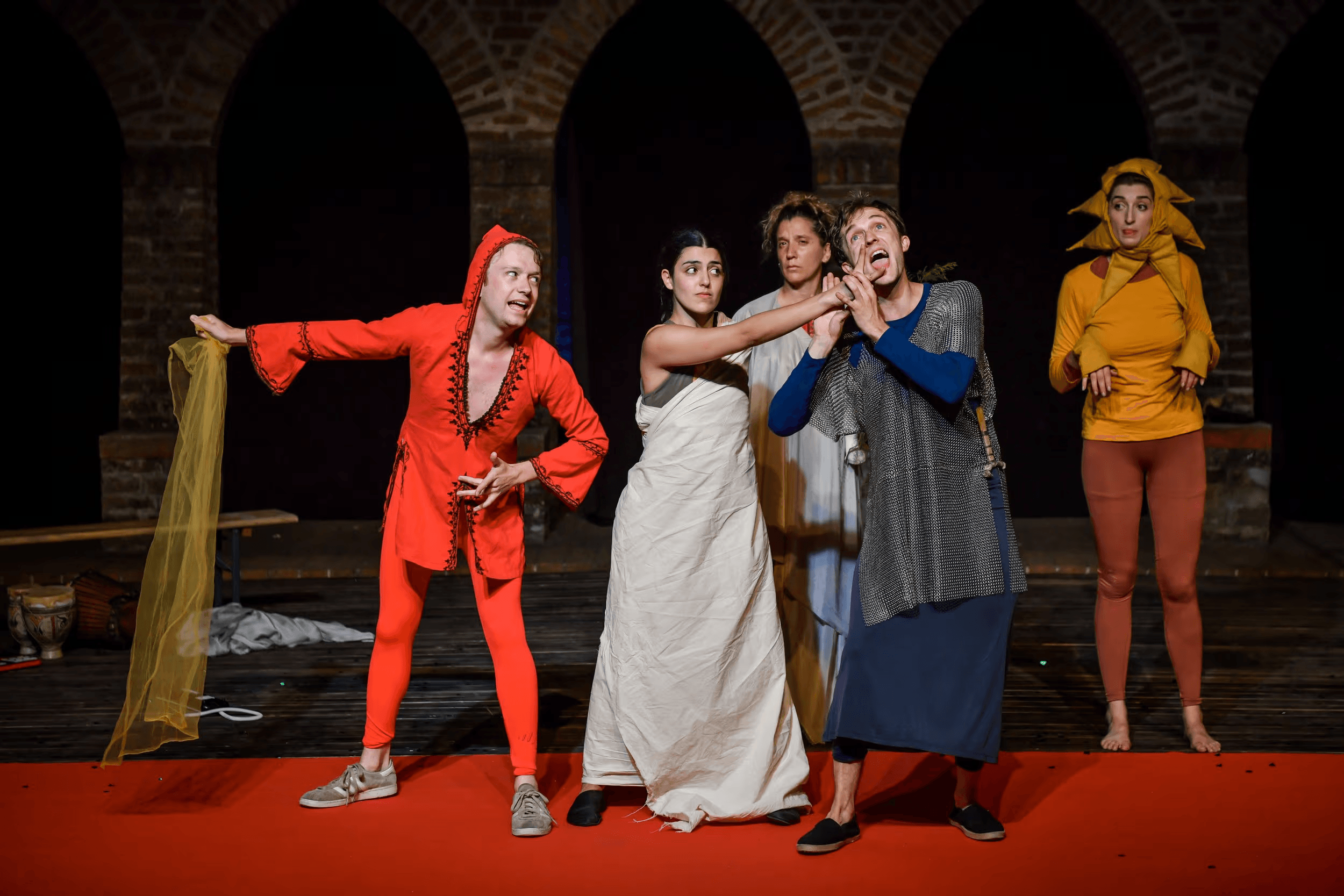 Actors performing a vibrant and expressive scene from a Flute Theatre production of Pericles, with colourful costumes and dynamic physical theatre, set against a backdrop of ancient stone arches.