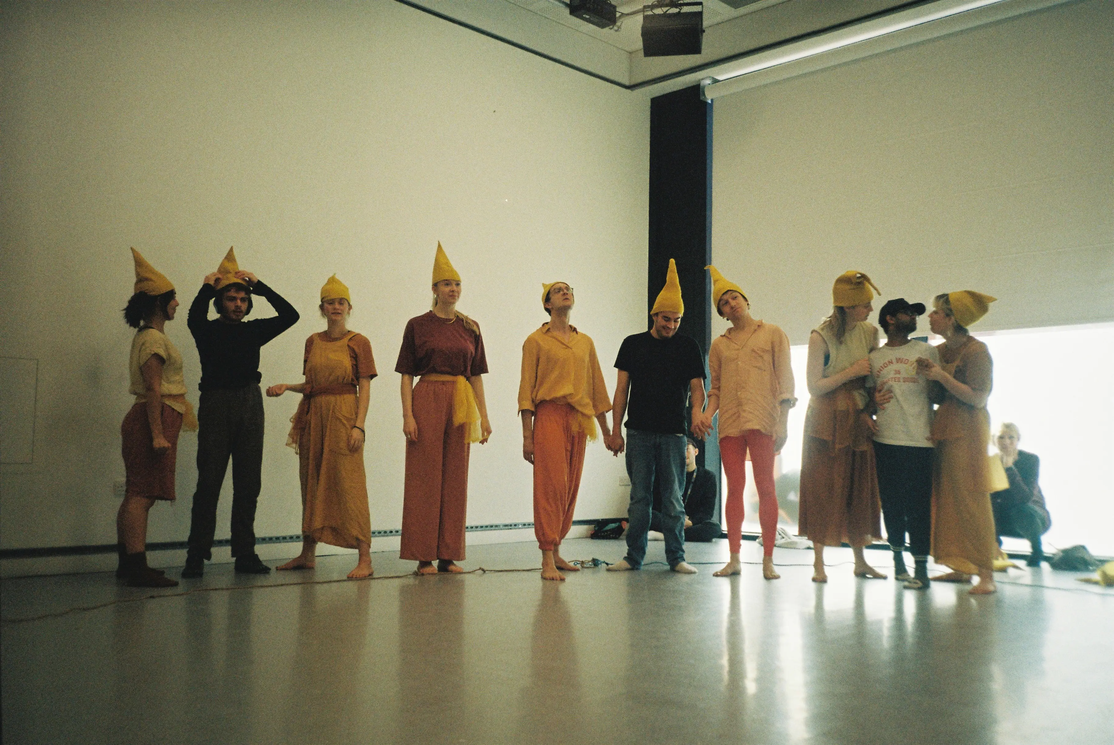 A group of performers in yellow and orange costumes with pointed hats stand in a line on stage, holding hands and looking upward during a Brighton inclusive theatre performance.