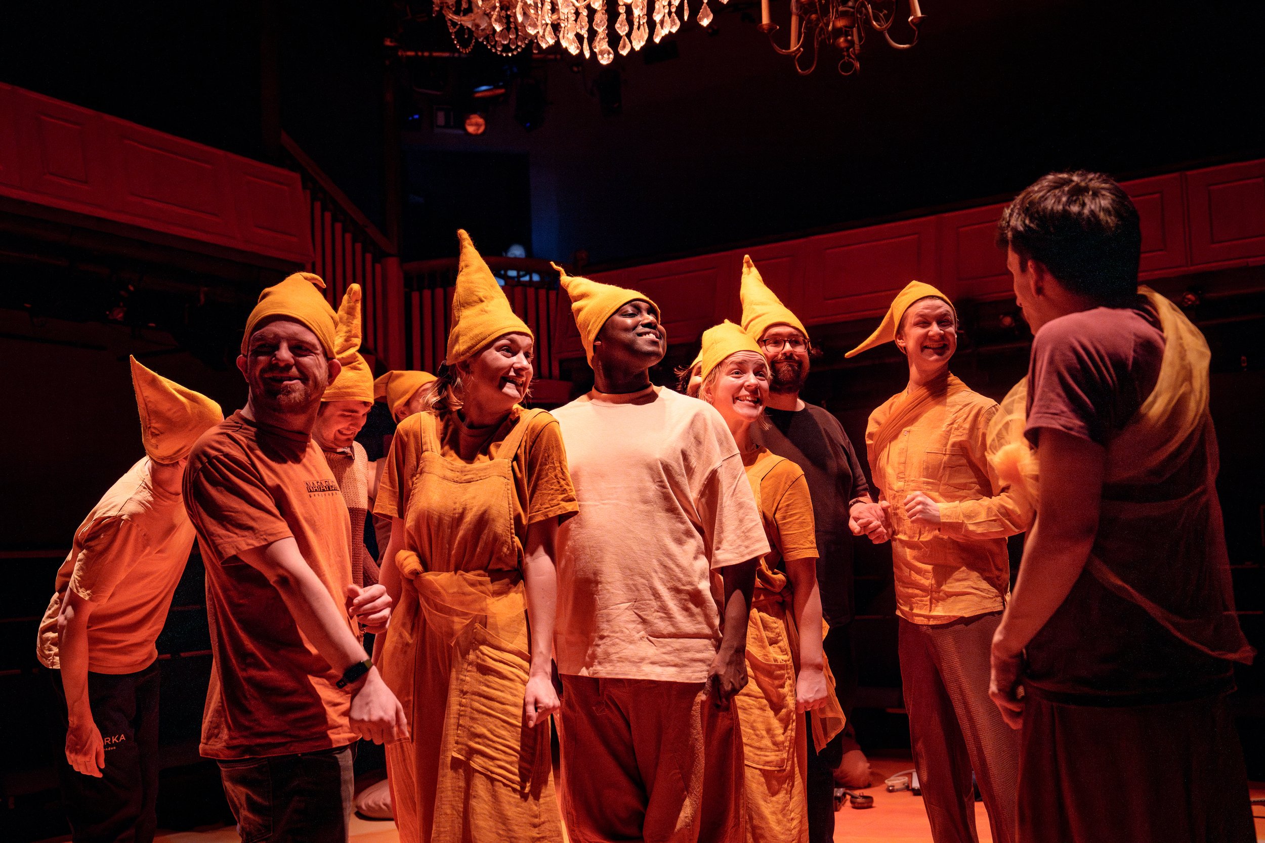A group of actors wearing orange and yellow costumes and pointy hats smile warmly during a live performance by Flute Theatre. The inclusive scene is lit with soft red stage lighting under a chandelier, showcasing Flute Theatre’s unique adaptation of Shakespeare for autistic individuals using the Hunter Heartbeat Method. The ensemble’s joyful expressions reflect the company’s commitment to sensory-friendly theatre experiences. Flute Theatre also offers training and workshops for actors, teachers, applied theatre students, and families to support neurodiverse communities.