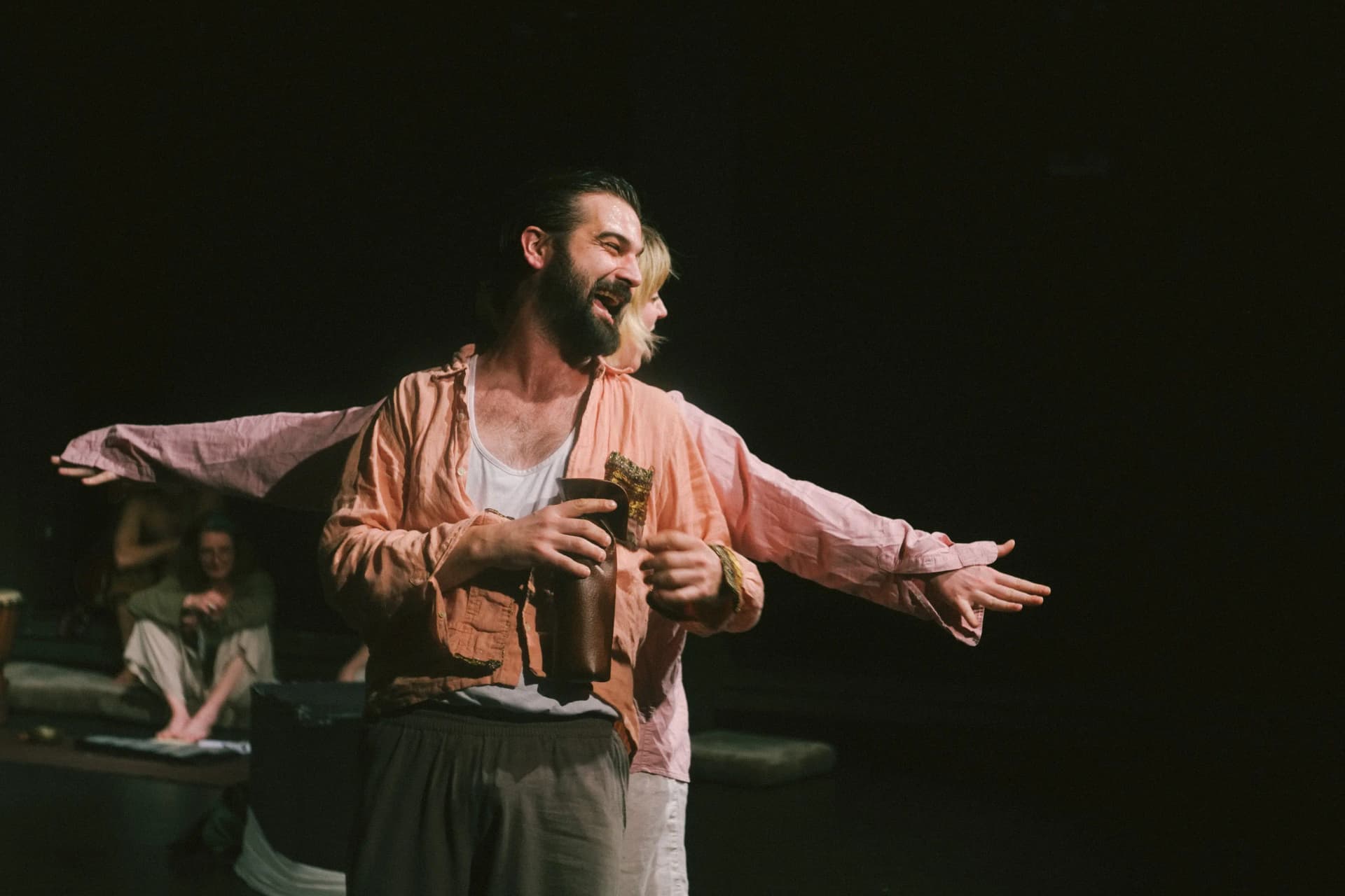 A bearded actor in an open shirt smiles broadly while holding a cup, with another performer behind him extending their arms playfully in a joyful moment on stage.