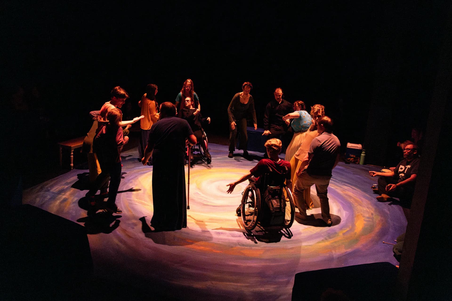 A group of diverse performers and audience members, including wheelchair users and children, stand in a circle on a stage painted with a swirling galaxy design, engaging in an inclusive performance.